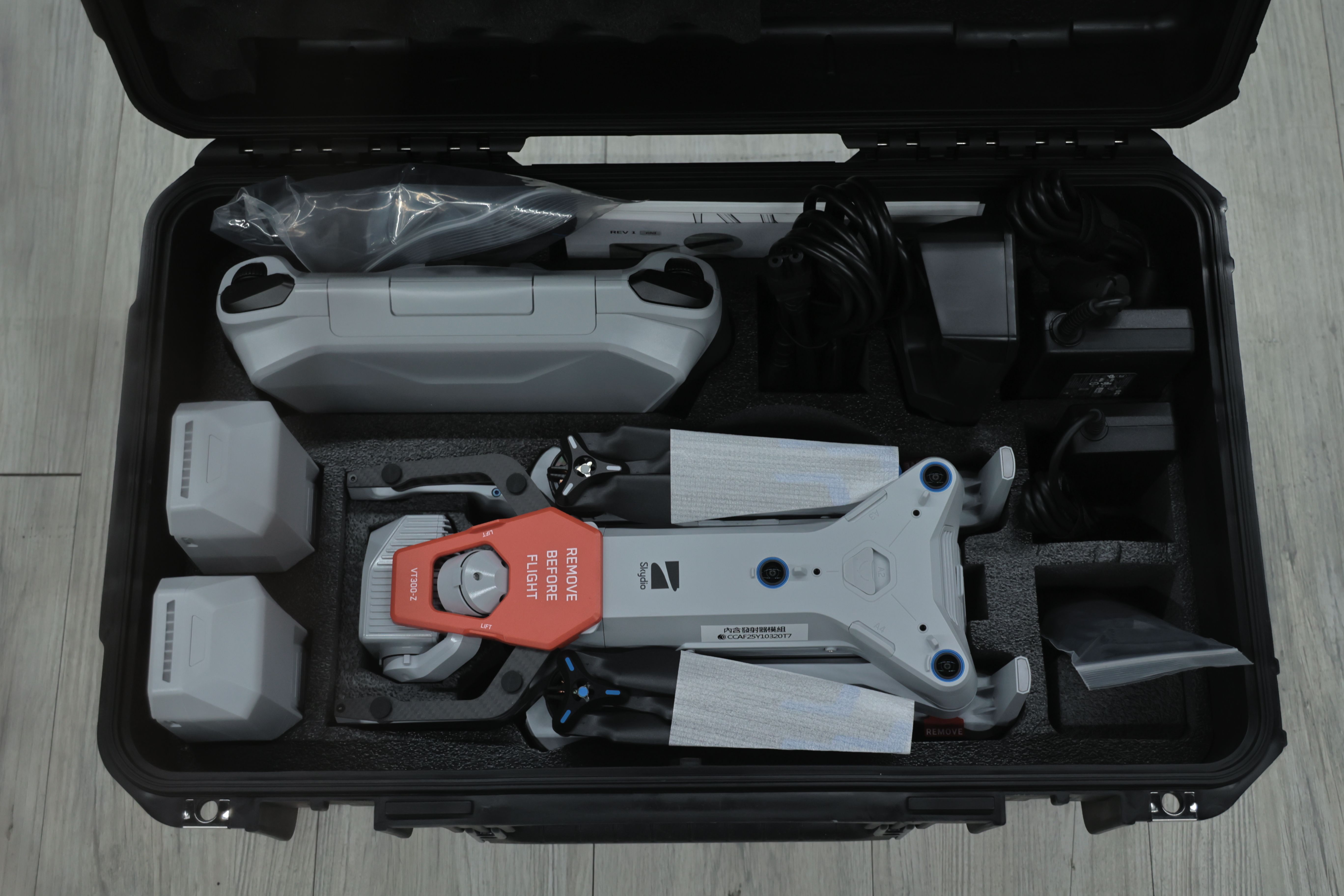 打開的黑色硬殼防護收納箱內部俯拍圖，Skydio X10 無人機置於中央泡棉槽位中，機身裝有紅色「REMOVE BEFORE FLIGHT」保護件，周圍可見兩顆電池、遙控器、充電設備、電源線與配件，整齊固定於箱內。