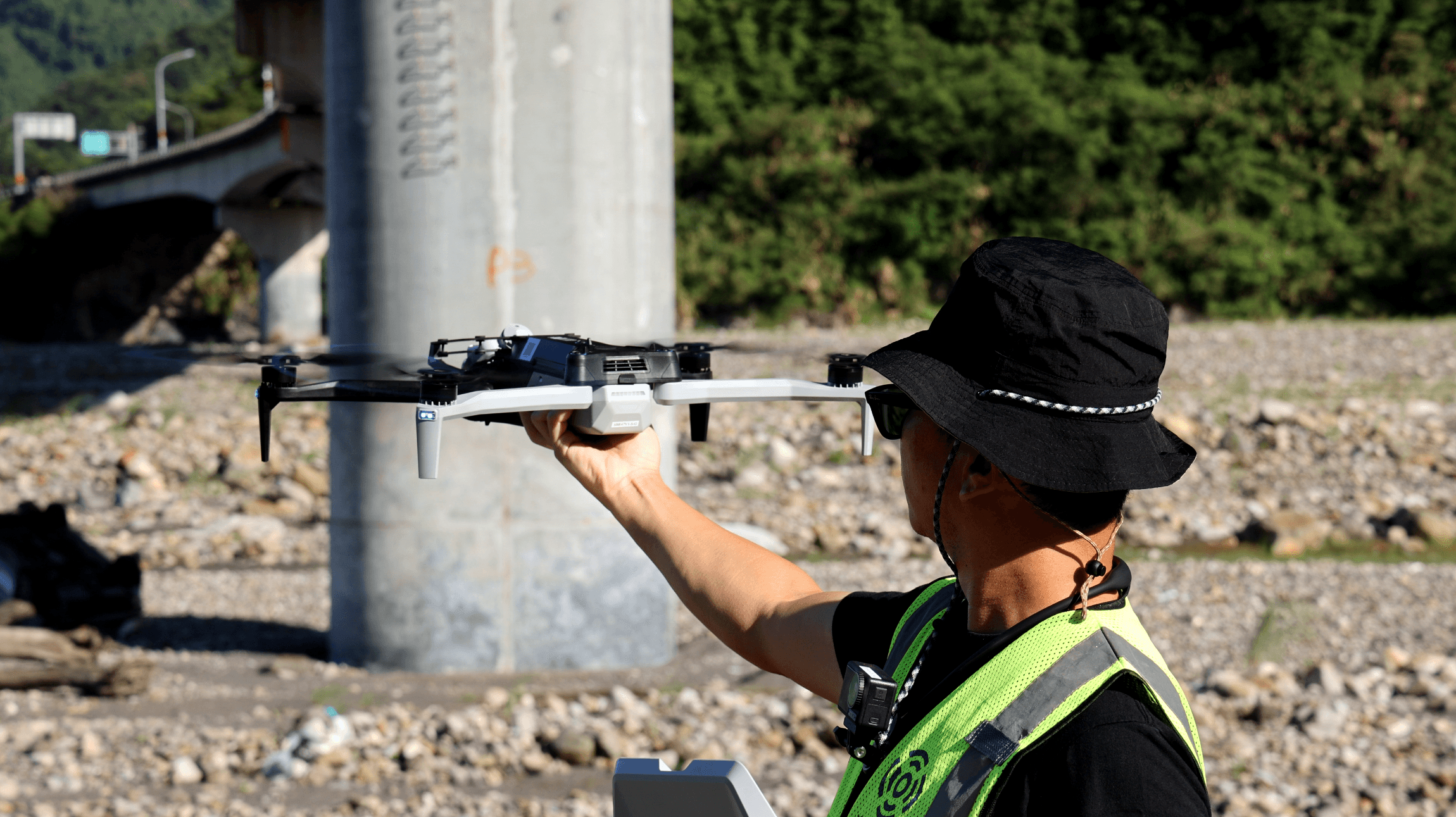 一名配戴安全帽與反光背心的作業人員在橋梁下方手持多旋翼無人機準備起飛,背景可見橋墩與河床環境,呈現無人機橋梁巡檢作業情境。